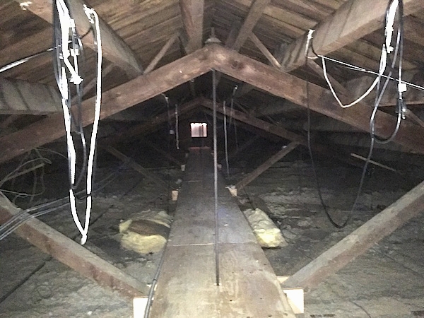 A view from across an attic with gray blown in insulation and yellow fiberglass batts with a cat walk leading to a gable vent at the end. There are wires hanging from the wood beams.
