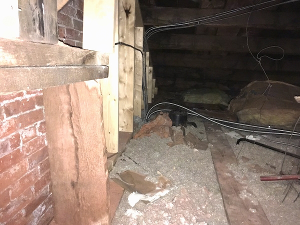 Gray blown in insulation next to a brick outer wall with wood beams and wires running around and under it.