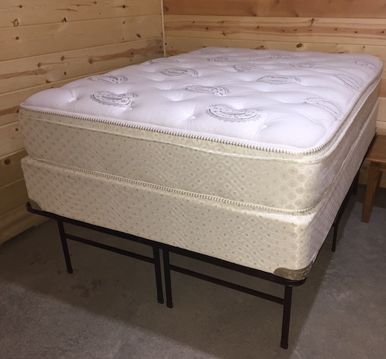 A white mattress and box-spring on a metal bed frame sitting in a room with wooden walls.