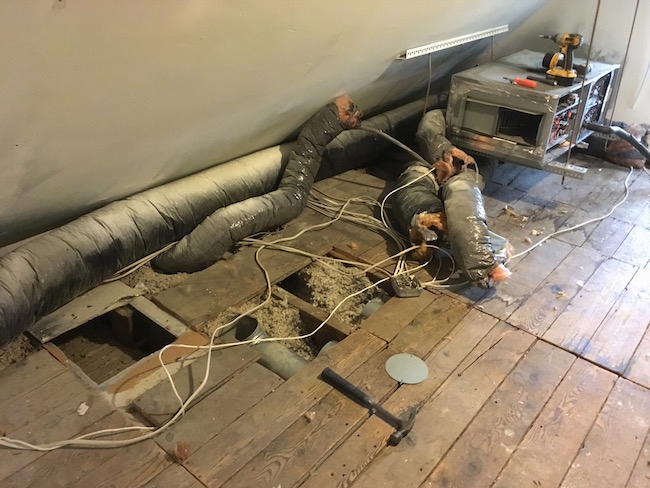 A view of an HVAC system taken apart with floor boards removed exposing the fiberglass under the wood floor with flex ducts laying on the floor