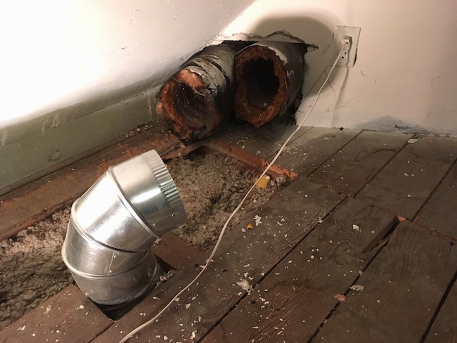 Flex ducts poking out from the wall in an attic with some boards removed with a silver duct also sticking out of the floor.
