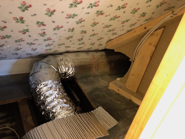 A metal duct wrapped in shiny bubble wrap connected to a foil duct that runs under the floor next to a wall with wall paper decorated in holly leaves