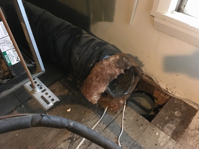 A large black flex duct with dirty orange fiberglass poking out the end laying disconnected from the wooden floor with parts of the HVAC system behind it.