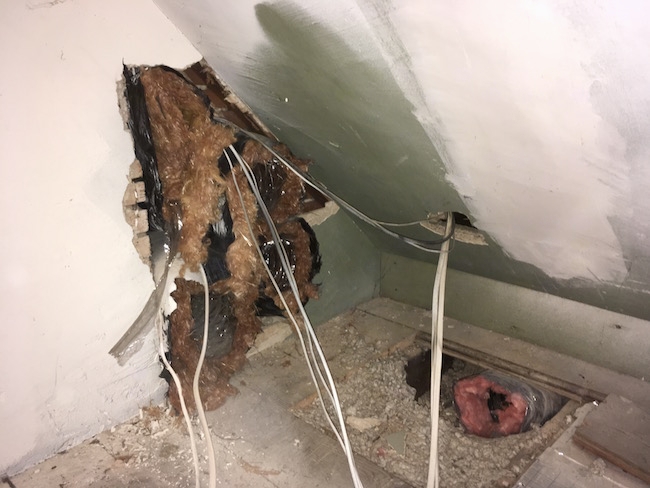 Black flex ducts coming out of a hole in the wall with brown-orange colored fiberglass hanging from it and a hole in the wooden floor showing gray blown in fiberglass and a black flex duct with pink fiberglass exposed at the end with white wires.