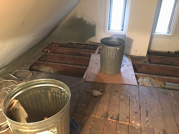 An attic floor with the boards removed towards the back of the room with two long skinny windows with the sun shining in and two metal trash cans and white wires. You can see the blown in gray fiberglass insulation between one of the grooves.