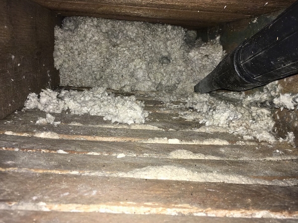 Inside of a wooden floor cavity half full with blown in loose white fiberglass insulation and a black shop vac tube vacuuming it out.