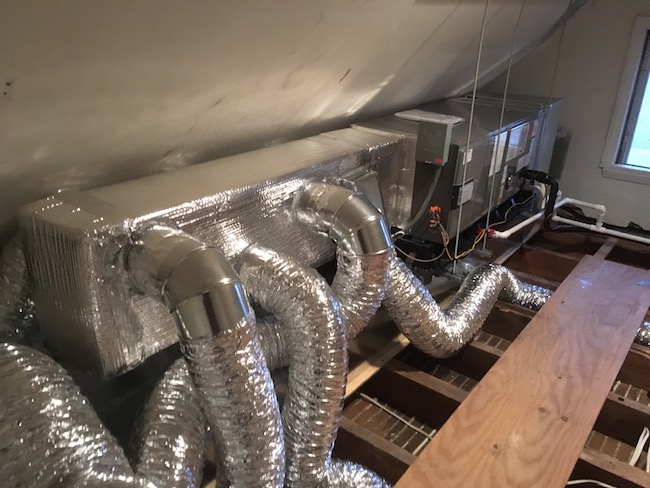An air handler wrapped in silver bubble wrap with foil ducts coming out of it with the wood floor removed showing the floor joists in an attic