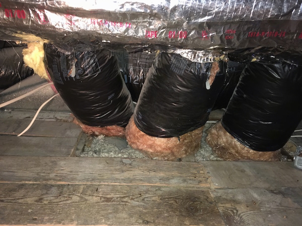 A hole in a wooden floor with large black flex ducts going down into the floor. There is orange fiberglass at the base of the ducts hanging out from the black plastic cover and loose gray fiberglass around the bottom of them on the floor with white wires off to the left and more exposed insulation.