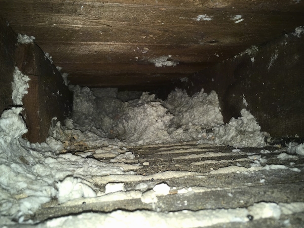 A view from inside of a floor cavity with white thick fiberglass clumped up in the back and scattered about with plaster showing in the cracks.