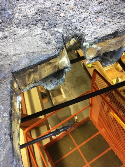 A hole cut in a flat roof where an HVAC unit was removed. The hole shows how the roof is mainly made of yellow fiberglass and there is a view of the side of the building below.