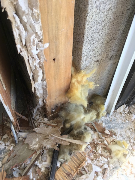 A crow bar in the end of a cinder block wall inside of a building pulling out clumps of moldy yellow fiberglass that is now sticking out from between the cinder blocks and the wood frame.