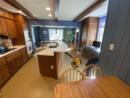 A man in a blue shirt holding a yellow and black Dewalt battery powered leaf blower up to the cracks between a countertop and the cabinets below