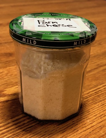 A glass jar with a green salsa lid with a white lable on it saying Parm Cheese with parmesan cheese inside sitting on a wooden table