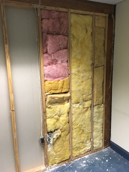 A wall with the drywall removed showing yellow and pink fiberglass between the beams.