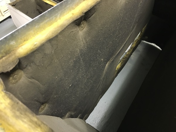 The inside of an air duct with old dusty yellow fiberglass lining the inside with a black dirty layer on top. The fiberglass is exposed around the edges and the fiberglass is dusty and dirty.
