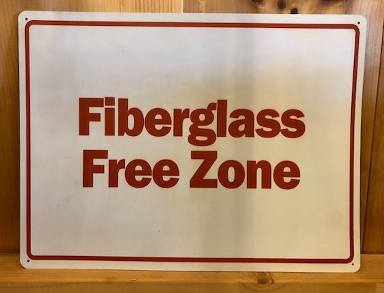 A white sign with red letters leaning against a wooden wall that reads Fiberglass Free Zone