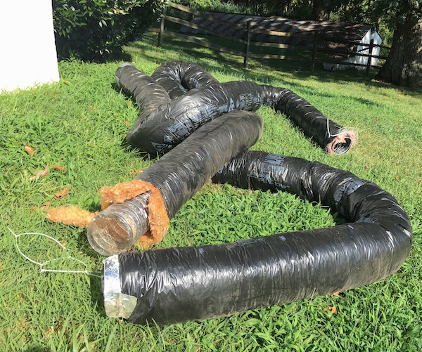 Long tubes of black flex ducts laying out in a grassy yard next to a house with an old spring house in the distance.