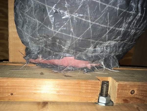 A gray flex duct with pink fiberglass insulation showing at the end where the duct meets the wood. There are white fiberlgass strings hanging out of the end. There is a screw and a nut holding the wood beam together.