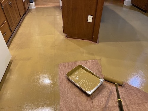 A kitchen floor part way painted with an olive-brown color with a paint tray and a roller