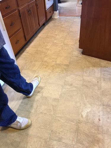 A man in blue jeans and white sneakers standing in a kitchen that has a tan linoleum floor.