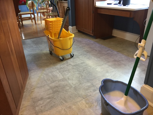Two mops and buckets, a yellow industrial size and a normal blue bucket on a linoleum floor in a kitchen
