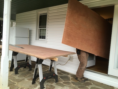 A man carrying a sheet of plywood into a white farm house with more wood on the porch