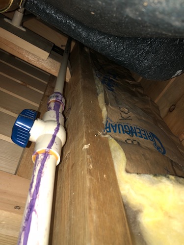 A white pipe running up a wooden wall attached to a wooden stud with a strip of yellow fiberglass batt insulation in the bay next to it.