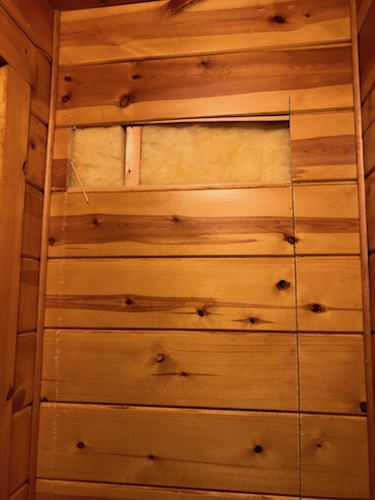 A rectangular hole in a wooden wall with yellow fiberglass batt insulation showing where teh wood was cut.