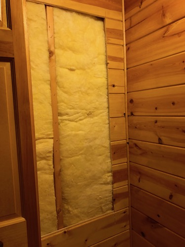 A large rectangular hole in a wooden wall with yellow fiberglass batt insulation exposed where the wood was cut away.