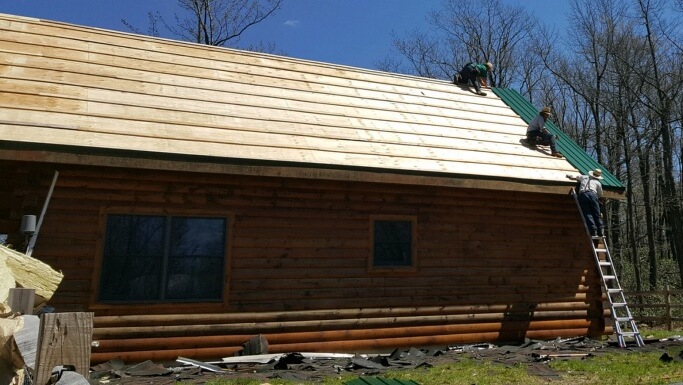 The side view of a log cabin roof with two men on the roof and one man on a ladder that is leaning against the side. There is a strip of green tin on the far right side of the roof. The rest is plywood.