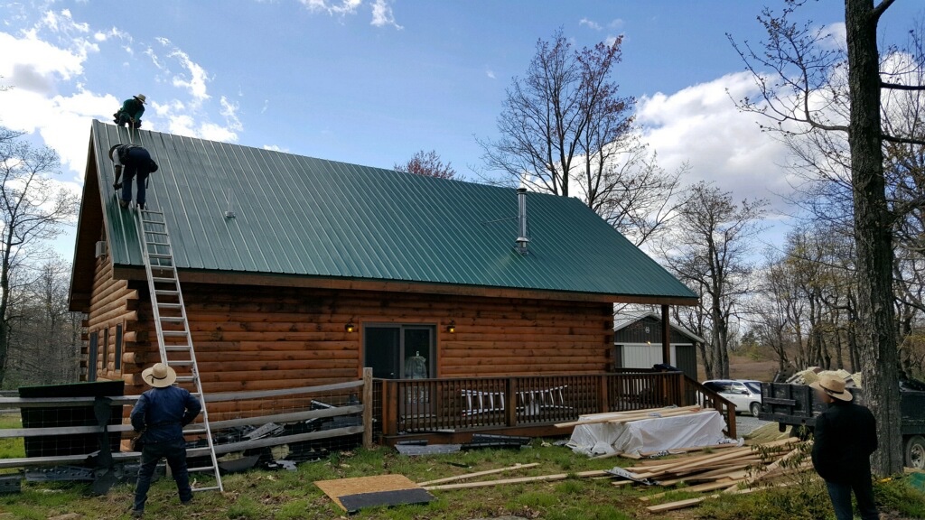 Back side view of a wooden log cabin with a green tin roof. There are two men on the roof. One is begining to come down the ladder and the other is up at the peak. Another man in a straw hat is standing on the ground looking up at the men on the roof. There are piles of wood and fiberglass in the yard and a fourth Amish man in a straw hat standing off to the right of the image.