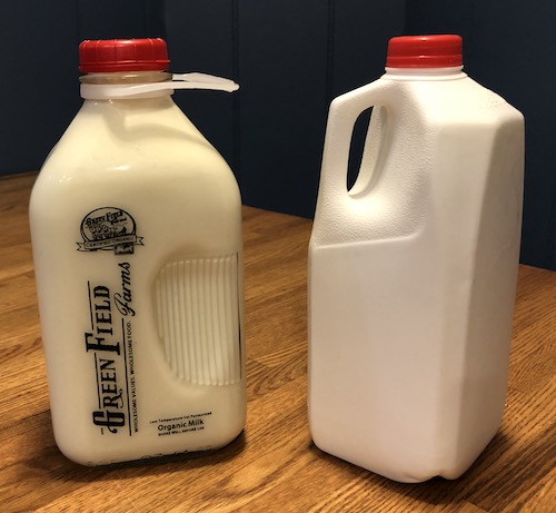 A glass container of grass fed milk next to a plastic milk jug on a wooden table