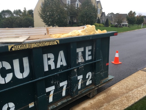 A dumpster full of wood and yellow fiberglass on the street in front of a house with an orange cone next to it
