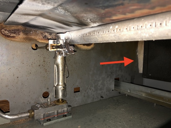 Inside of a gas oven next to the pilot light is a red arrow pointing to the right at exposed ceramic fiber wool.