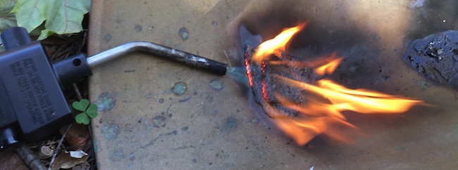 A blow torch blowing flames over top of a roof shingle on a copper surface.