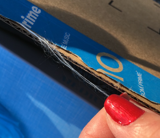 A brown cardboard box with blue packing tape across the top with strands of white glass coming from under the ripped tape. There is a hand with red painted fingernails holding the end of the fiberglass strand.