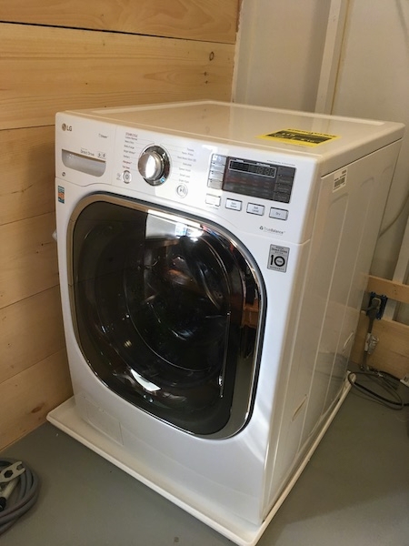 A white ventless washer dryer combo sitting on a white plastic tray on top of a gray floor next to a wood wall.