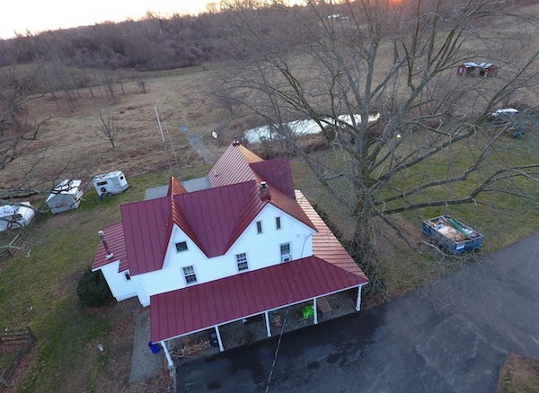 An aerial view of a white farm house with a red tin roof. It has a wrap around porch. There is a pond behind the house a dumpster some sheds, a horse trailer and a couple of lean-tos in the field behind the house.