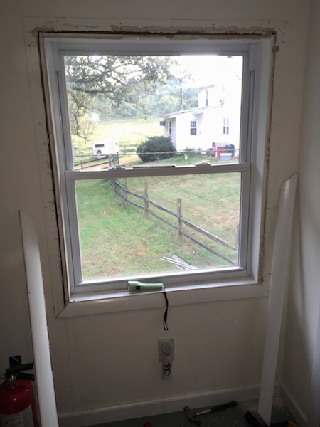 A window with the wood frame removed to expose the crack around the window. There is a view of the white farm house outside.