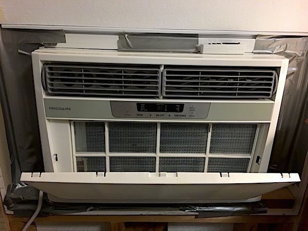 A white Frigidaire window heat pump unit inside of a window with the front panel open and duct tape around the edges of the window sill.