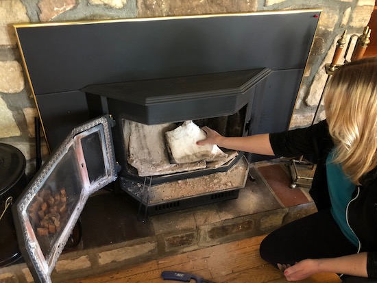 A person kneeling down in front of a wood burning stove with her hand on a white bat of ceramic insulation that was removed from the top of the stove.