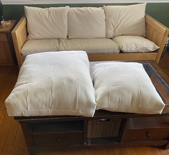 Two white cushions sitting on a coffee table in front of a wooden This End Up couch that has chemical-free organic replacement cushions on it